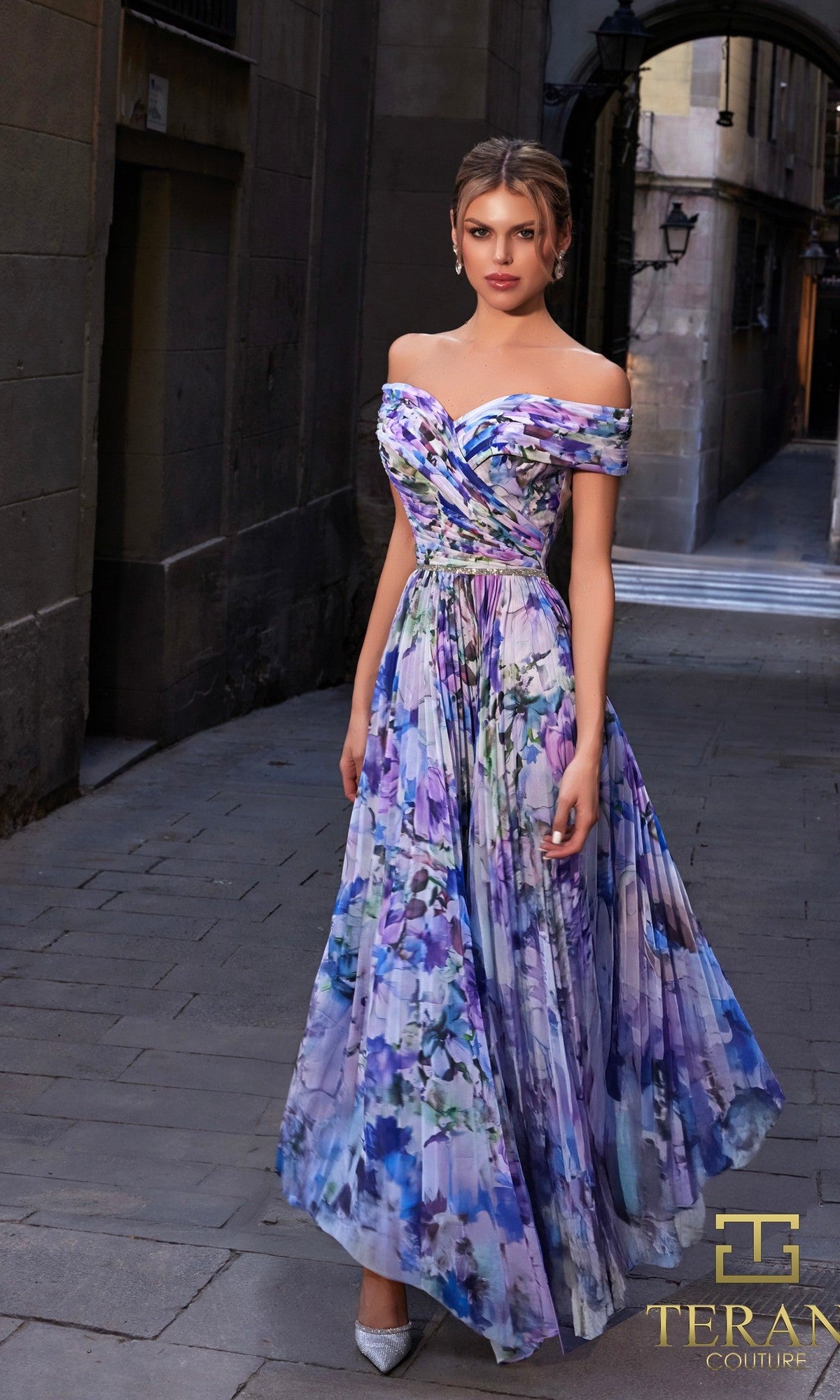 A woman wearing the Long Formal Dress: Terani 261C6641 stands on a narrow stone street, looking at the camera in silver shoes, with the